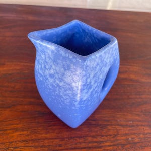 May include: A blue ceramic pitcher with a unique, angular design. The pitcher has a handle and a spout, and the surface is speckled with white. The pitcher is sitting on a wooden surface.