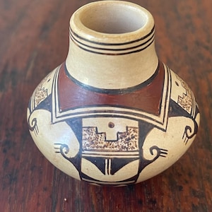 May include: A small, round ceramic vase with a cream-colored base, featuring hand-painted geometric patterns in dark brown and deep red. The design includes intricate black lines, stippled textures, and stylized motifs around the body.