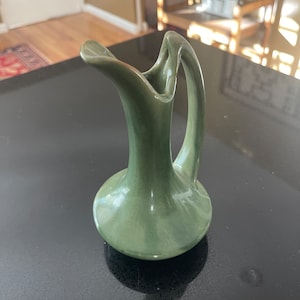 May include: A green ceramic pitcher with a unique, curved handle. The pitcher has a smooth, glazed finish and a slightly textured surface.