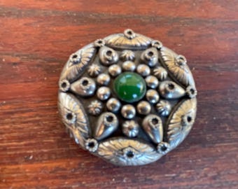 May include: A round, silver-toned brooch with a central green stone. The brooch features a detailed design of small silver spheres and petal-shaped elements. The brooch is set against a wooden surface.