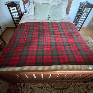 May include: A rectangular blanket featuring a dark red and forest green plaid pattern. The fabric has a soft, textured appearance and is draped across a bed, covering the surface from the headboard to the footboard with a consistent tartan design.