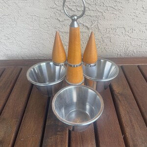 May include: A stainless steel condiment server with a wooden handle and three conical wooden toppers. The server has four stainless steel bowls. The handle has a silver ring at the top.