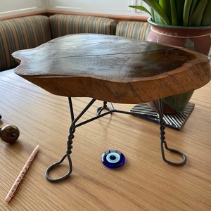 May include: A wooden table top with a natural edge sits on a black metal stand. The table top is dark brown and has a natural wood grain. The stand is made of twisted wire and has three legs. A blue evil eye charm is on the table top.