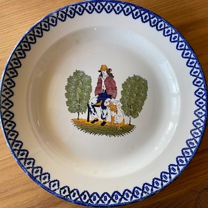 May include: A white ceramic plate with a blue and white border design. The center of the plate features a hand-painted scene of a person sitting with a dog in a forest setting.