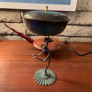 May include: A vintage mid-century modern style table lamp with a stylized bronze sculpture of a person holding up a blue and green ceramic bowl with a lid. The lamp is on a wooden table.