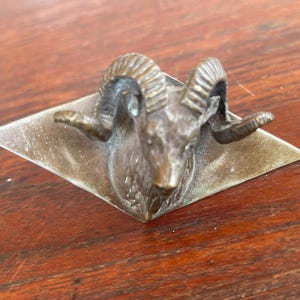May include: A bronze ram head sculpture mounted on a diamond-shaped base. The ram has detailed horns and facial features. The sculpture is set against a dark wood surface, creating a contrast in color and texture.