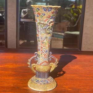 May include: A colorful, ornate vase with a flared top and two handles. The vase features intricate patterns in red, blue, yellow, and green. The base is white with a gold spiral design, and the top rim is gold.
