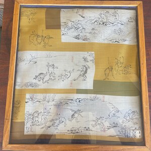 May include: A framed textile piece featuring a mustard yellow and olive green background with three horizontal white panels. The panels contain black ink illustrations of anthropomorphic rabbits and frogs engaged in various activities.