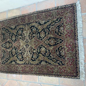 May include: Rectangular rug with a detailed floral pattern in black, gold, and burgundy. The rug has a burgundy border and white fringe on the short ends. The rug is placed on a brick surface.