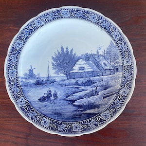 May include: A blue and white decorative plate with a rural landscape scene. The plate features a village scene with a windmill, boat, and figures near a river. The rim has a floral and scroll design.