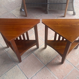 May include: Two matching triangular side tables crafted from medium-toned wood. Each table features a flat, smooth top surface, three sturdy legs, and a lower triangular shelf supported by vertical slats connecting the shelf to the tabletop frame.