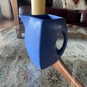 May include: A blue ceramic pitcher with a unique, angular design. The pitcher has a large, rounded handle and a spout. The color is a solid, matte blue. The pitcher is sitting on a wooden surface.