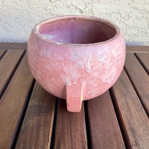 May include: A round, pink ceramic planter with a textured surface and a small, rectangular handle. The planter has a wide opening and a faded, aged appearance. The interior is a darker shade of pink. It sits on a wooden surface.