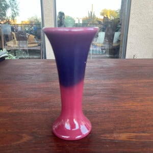 May include: A two-toned vase with a flared top and a rounded base. The top half is a deep purple, transitioning to a vibrant pink at the base. The vase is made of a glossy material and sits on a wooden surface.