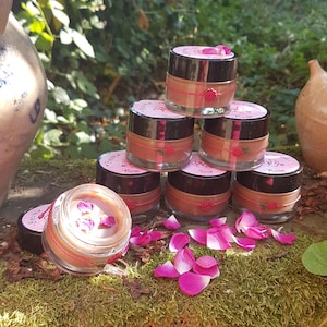 Peut inclure: Une pile de petits pots en verre transparent avec des couvercles noirs contenant une crème brun clair. Chaque pot est entouré d'une ficelle rouge avec une petite breloque rose rouge. Des pétales de rose rose sont éparpillés à la base.