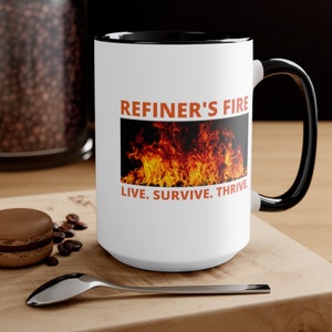 May include: White ceramic mug with a black interior and handle. The mug features the text "REFINER'S FIRE" above a graphic of flames, with the words "LIVE. SURVIVE. THRIVE." below. The mug is on a wooden surface.