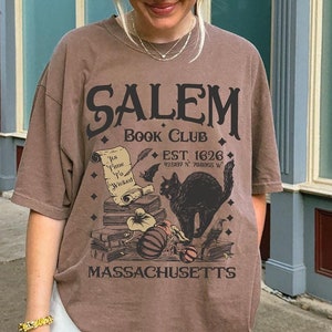 May include: A brown t-shirt with the text "Salem Book Club" and a graphic of a black cat, books, and a pumpkin. The shirt also includes the text "Est. 1626" and "Massachusetts". The shirt is worn with white pants.