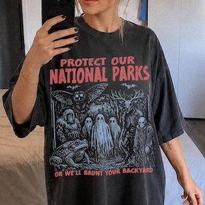 May include: A charcoal grey oversized t-shirt featuring the red text "PROTECT OUR NATIONAL PARKS" at the top and "OR WE'LL HAUNT YOUR BACKYARD" at the bottom. The graphic depicts a dark forest scene with an owl, a toad, ghosts, and a skeletal figure.