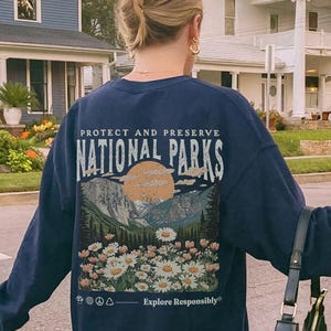 Op de afbeelding: Een marineblauw sweatshirt met lange mouwen en een afbeelding op de achterkant met de tekst "PROTECT AND PRESERVE NATIONAL PARKS" boven een illustratie van bergen, een zon en madeliefjes, met de tekst "Explore Responsibly" eronder.