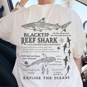May include: Cream-coloured t-shirt featuring a detailed black and white illustration of a Blacktip Reef Shark, with accompanying text. The shirt includes scientific details and the words "EXPLORE THE OCEANS".