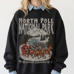 Op de afbeelding: Zwart sweatshirt met een afbeelding van de Kerstman in een slee getrokken door rendieren, met de tekst "NORTH POLE NATIONAL PARK" en "EXPLORE RESPONSIBLY". Het ontwerp toont een bergketen, bomen en een rivier.