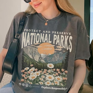 May include: A gray t-shirt with a graphic of a mountain landscape, a setting sun, and daisies, with the text "Protect and Preserve National Parks." The shirt also has the text "Explore Responsibly."