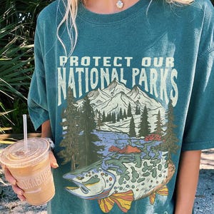 May include: A teal green t-shirt with a graphic print of a mountain scene and a fish. The text on the shirt reads "Protect Our National Parks" and "Explore Responsibly".
