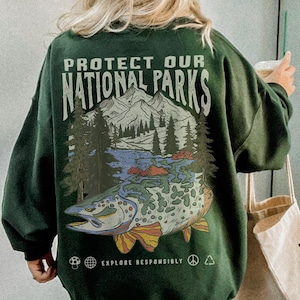 以下が含まれることがあります： 「PROTECT OUR NATIONAL PARKS」の文字と、魚、山、木々のグラフィックが描かれた濃い緑色のスウェットシャツ。スウェットシャツには「EXPLORE RESPONSIBLY」の文字とその他のシンボルも描かれています。スウェットシャツが着用されています。