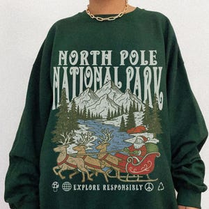 May include: Dark green sweatshirt with a graphic of Santa Claus in a sleigh pulled by reindeer, set against a mountain and lake scene. The text "NORTH POLE NATIONAL PARK" and "EXPLORE RESPONSIBLY" are printed on the front.