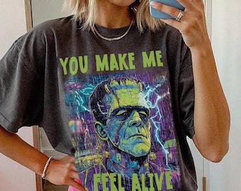 Puede incluir: Camiseta gris oscuro con un gráfico de Frankenstein y el texto "YOU MAKE ME FEEL ALIVE". Se combina con shorts rosa brillante. La persona lleva un gorro azul y gafas de sol.