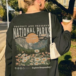 Op de afbeelding: Een zwart sweatshirt met ronde hals en een rugprint met de tekst "PROTECT AND PRESERVE NATIONAL PARKS" boven een illustratie van bergen, een zon en een veld met madeliefjes, met de tekst "Explore Responsibly" en kleine iconen eronder.