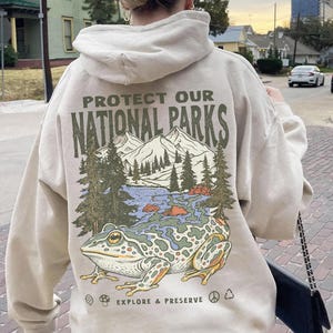 May include: A beige hooded sweatshirt featuring a graphic on the back with the text PROTECT OUR NATIONAL PARKS and EXPLORE & PRESERVE. The illustration depicts a large frog in the foreground with a mountain, forest, and lake scene behind it.