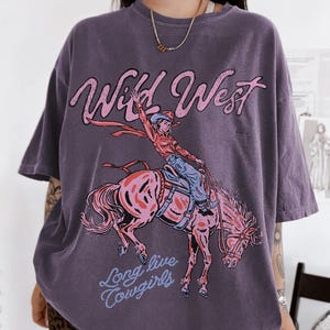May include: A dark purple short-sleeve t-shirt featuring a graphic print of a person riding a bucking horse. The text Wild West is printed in pink above the graphic, and the text Long live cowgirls is printed in light blue below the horse.
