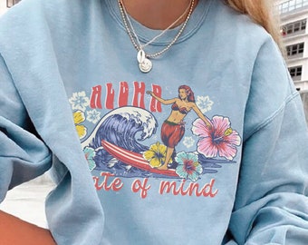 May include: A light blue sweatshirt featuring a colorful graphic of a surfer girl, waves, and tropical flowers. The text "Aloha State of Mind" is printed above and below the image in red and white. The sweatshirt has long sleeves and a crew neck.