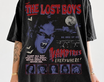 May include: A black short-sleeve t-shirt featuring a graphic print from the movie The Lost Boys. The design includes red and blue text, a large vampire face, bats, a moon, and five character portraits with the phrase Sleep All Day Party All Night.