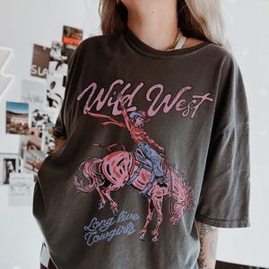 Puede incluir: Una camiseta gris carb&oacute;n de corte oversize con un estampado gr&aacute;fico de una vaquera montando un caballo encabritado. El texto "Wild West" aparece en rosa sobre la ilustraci&oacute;n, y "Long live cowgirls" est&aacute; impreso en azul bajo el caballo.