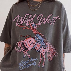 May include: A charcoal gray short-sleeve t-shirt featuring a graphic print of a person riding a bucking horse. The text "Wild West" appears in pink above the illustration, and the text "Long live cowgirls" appears in light blue below it.