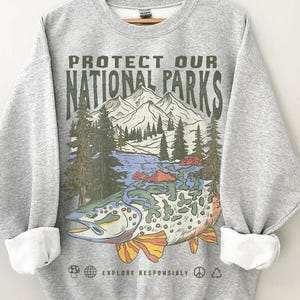 Puede incluir: Una sudadera gris con mangas dobladas que presenta un gr&aacute;fico de un pez con una escena de monta&ntilde;a y lago. El texto "PROTECT OUR NATIONAL PARKS" est&aacute; impreso encima de la imagen, con "EXPLORE RESPONSIBLY" debajo.