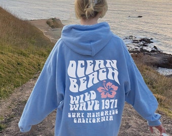 May include: A light blue hooded sweatshirt with a graphic print on the back that says "Ocean Beach Wild Wave 1971 Surf Memories California". The graphic includes a pink hibiscus flower. The sweatshirt is shown on a person standing on a path overlooking the ocean.