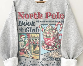 May include: Ash gray sweatshirt with a graphic that reads "North Pole Book Club." The graphic features a woman reading by a fireplace and a cup of hot cocoa with marshmallows and a candy cane. The text "Magical Reads" and "Hot Cocoa Served daily" are also included.