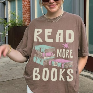 Puede incluir: Camiseta marrón con el texto blanco "READ MORE BOOKS". El gráfico presenta una pila de libros en verde azulado y rosa. La camiseta se usa con pantalones cargo blancos y gafas de sol.