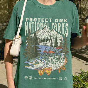 May include: A light green t-shirt with a graphic print of a mountain scene and a fish. The text "Protect Our National Parks" is printed on the shirt. The shirt also has the text "Explore Responsibly" printed on it.