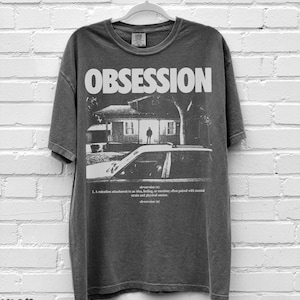 May include: A charcoal gray short-sleeve t-shirt featuring the word OBSESSION in large white letters above a black and white graphic of a house and a car. Below the graphic, the text defines obsession as a relentless attachment to an idea.