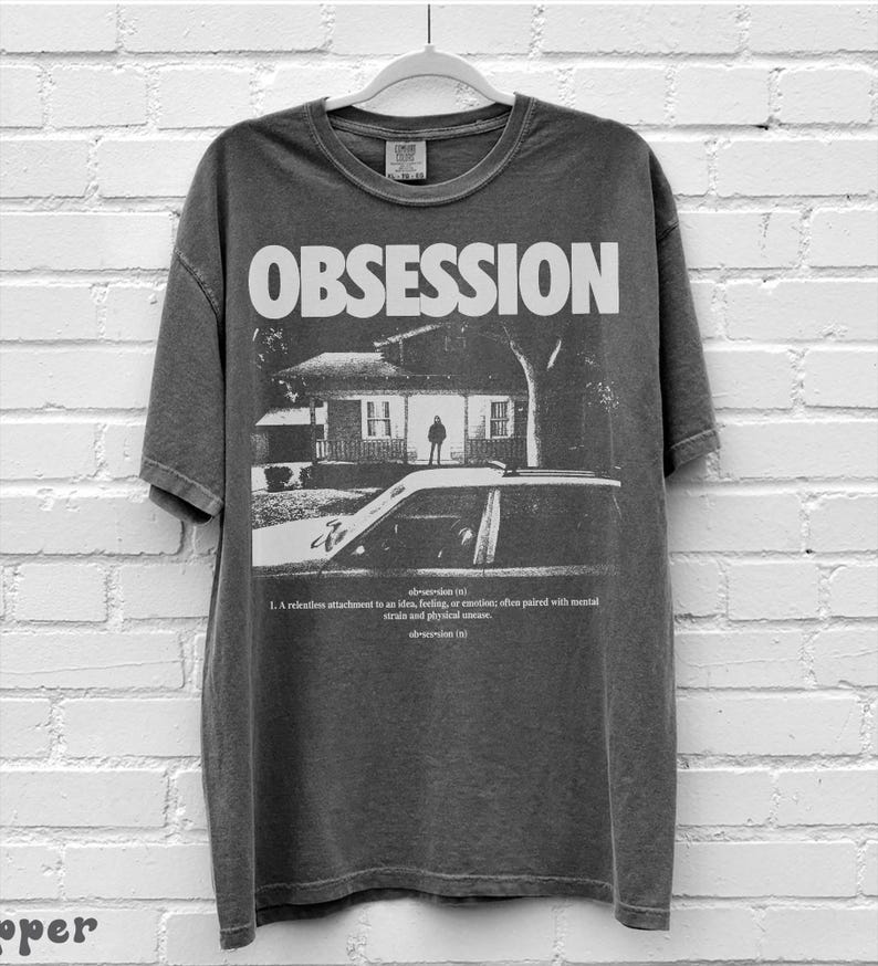 May include: A charcoal gray short-sleeve t-shirt featuring the word OBSESSION in large white letters above a black and white graphic of a house and a car. Below the graphic, the text defines obsession as a relentless attachment to an idea.