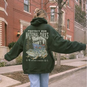 May include: A dark green hooded sweatshirt featuring a graphic on the back with the text "PROTECT OUR NATIONAL PARKS" and "EXPLORE & PRESERVE". The illustration depicts a large frog in the foreground with mountains, trees, and a river.
