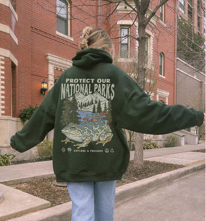 May include: A dark green hooded sweatshirt featuring a graphic on the back with the text "PROTECT OUR NATIONAL PARKS" and "EXPLORE & PRESERVE". The illustration depicts a large frog in the foreground with mountains, trees, and a river.