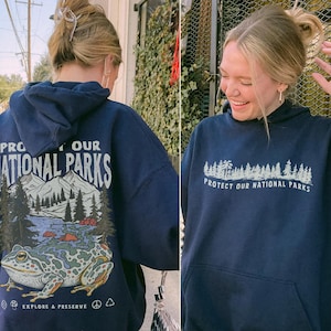 May include: A navy blue hooded sweatshirt featuring a graphic on the back of a large frog, mountains, and trees with the text "PROTECT OUR NATIONAL PARKS" and "EXPLORE & PRESERVE". The front displays a smaller forest graphic and the same text.