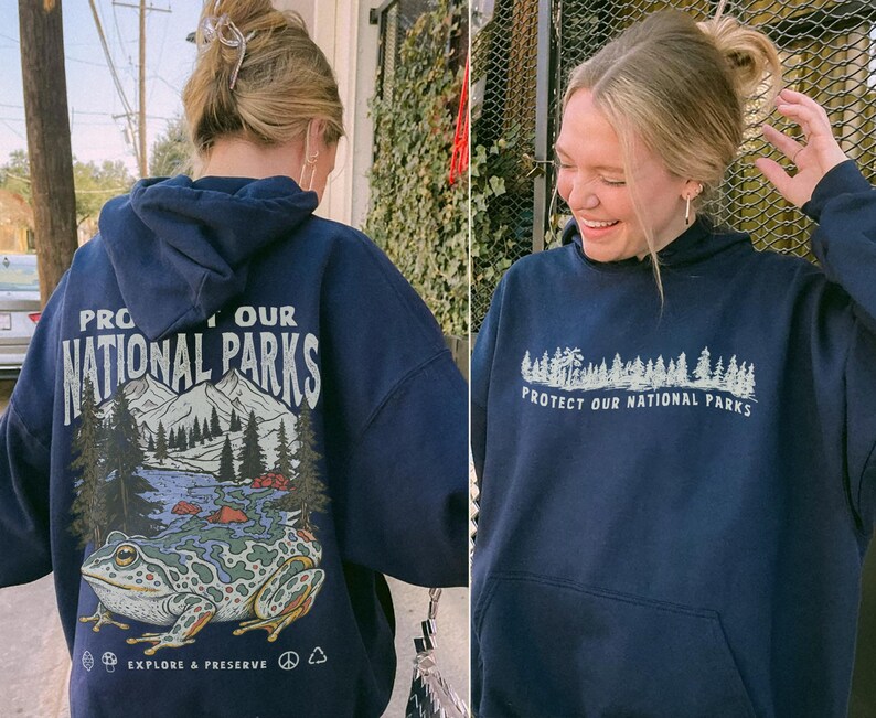 May include: A navy blue hooded sweatshirt featuring a graphic on the back of a large frog, mountains, and trees with the text "PROTECT OUR NATIONAL PARKS" and "EXPLORE & PRESERVE". The front displays a smaller forest graphic and the same text.