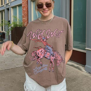 Puede incluir: Una camiseta marr&oacute;n de manga corta con un estampado gr&aacute;fico de una persona montando un caballo que corcovea. El texto Wild West est&aacute; impreso en rosa sobre el gr&aacute;fico y el texto Long live Cowgirls en azul claro debajo del dise&ntilde;o.