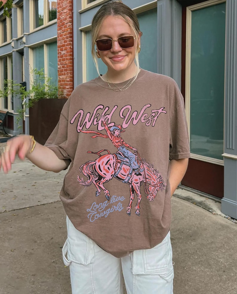 Puede incluir: Una camiseta marr&oacute;n de manga corta con un estampado gr&aacute;fico de una persona montando un caballo que corcovea. El texto Wild West est&aacute; impreso en rosa sobre el gr&aacute;fico y el texto Long live Cowgirls en azul claro debajo del dise&ntilde;o.